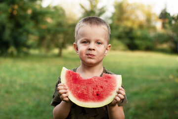 a fair-haired little boy holds a large piece of watermelon in his hands. a kid in the garden eats a watermelon
