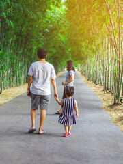 Fototapeta na wymiar Back view of Asian father hand holding lovely daughter walking on pathway through bamboo garden.Dad and daughter walking together in park. Happy family spending time together outside in green nature.