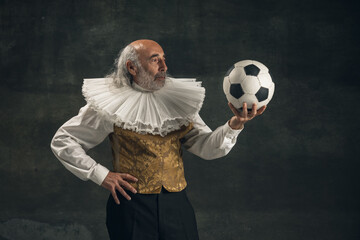 Elderly gray-haired man, actor posing with football ball isolated on dark vintage background. Retro...
