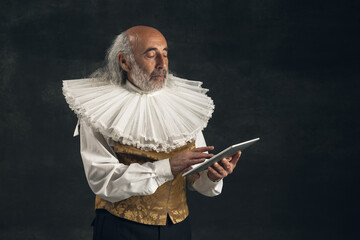 Portrait of elderly gray-haired man, medieval hystorical person, actor using tablet isolated on dark vintage background. Retro style, comparison of eras concept.