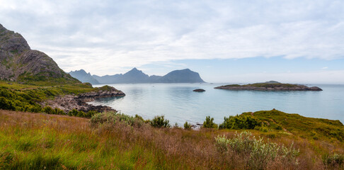 Picturesque grass covered rocky bay of the Norwegian fjord overlooking the distant mountains that are reflected in the surface of the sea under the blue sky.