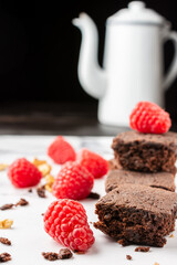 Close-up of chocolate brownie with crumbs, walnuts, red raspberries and white coffee pot, selective focus, on white marble table, black background, vertical