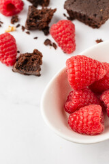Top view of white bowl with raspberries, pieces of chocolate brownie and walnuts, selective focus, on white table, vertical, with copy space