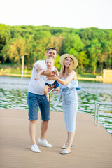 Fototapeta premium happy young parents mom and dad with their baby son walk in the summer by the water by the sea on the embankment and have fun, throwing the child up like a plane