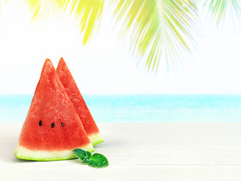 Two triangular slices of watermelon and mint on table top on blurred background of blue sea beach and palm tree at hot sunny day. Stand counter for product display or visual layout design.