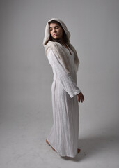 Full length portrait of young woman wearing classical white gown and a head covering veil in biblical style, sitting pose on light studio background.