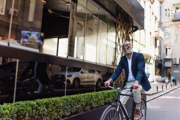 happy middle aged man in blazer and glasses riding bicycle on modern urban street