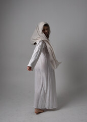 Full length portrait of young woman wearing classical white gown and a head covering veil in biblical style, sitting pose on light studio background.