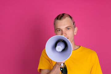 European handsome young man in yellow t shirt on pink background shouting in megaphone