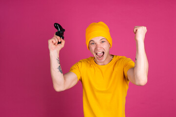 European handsome young man in yellow t shirt on pink background gamer with joystick  happy  shows winner gesture