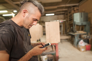 Carpenter artisan resting in workshop and scrolling on smartphone. Handsome handicraftsman texting message and tapping on phone