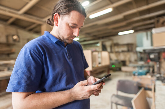 Carpenter artisan resting in workshop and scrolling on smartphone. Handsome handicraftsman texting message and tapping on phone