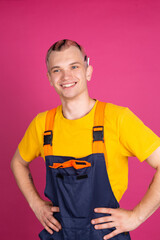 European handsome young man in yellow t shirt and overalls on pink background smiles  hands on waist cigarette  behind ear