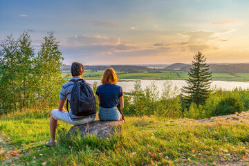 Rear view of couple sitting on hill against beautiful summer landscape with forest and lake in summer evening at sunset. Tourists man and woman admiring wonderful northern nature. Vacation concept