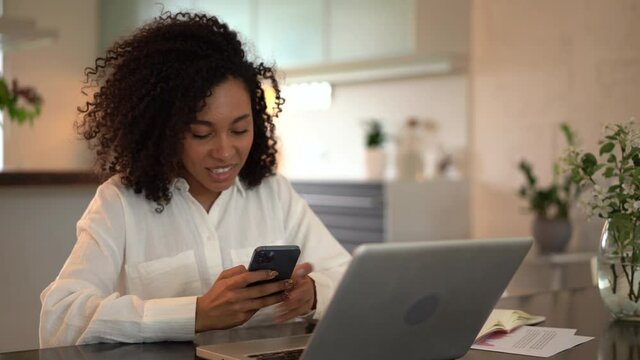 Joyful young mixed race woman chatting use smartphone enjoying break during work in front of laptop. Smiling female typing message or texting on mobile sitting on desk at home office