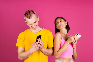 Beautiful modern young couple friends man and woman on pink background hold mobile phones girlfriend peeks at boyfriends screen