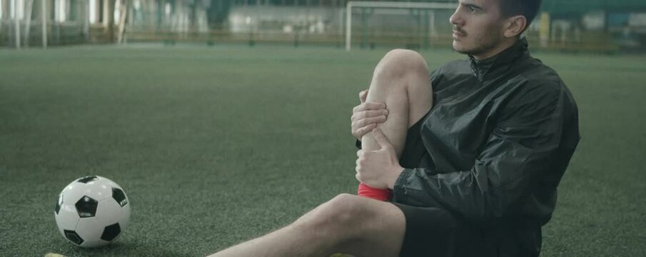 Anamorphic tilt up shot of Caucasian football player in sportswear sitting on indoor soccer field and pressing knees to chest while stretching hips before workout