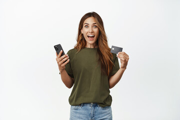 Fototapeta premium Online shopping. Excited smiling middle aged woman, holding smartphone, showing credit discount card, talking about big sale, standing against white background