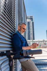 mature man in sunglasses using smartphone near blurred e-scooter