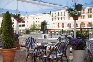 Summer cafe on the waterfront in Rosa Khutor, Sochi: tables and wicker chairs among decorative plants in the foreground.