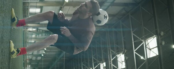 Vertical anamorphic shot of young Caucasian athlete juggling soccer ball while having workout on indoor field - Powered by Adobe