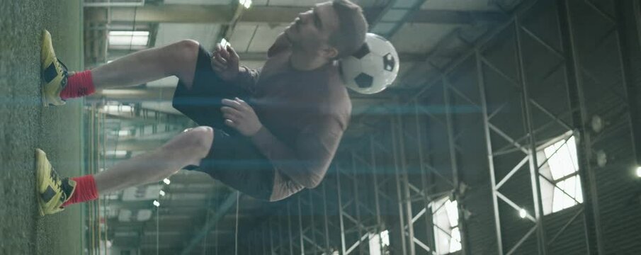 Vertical anamorphic shot of young Caucasian athlete juggling soccer ball while having workout on indoor field