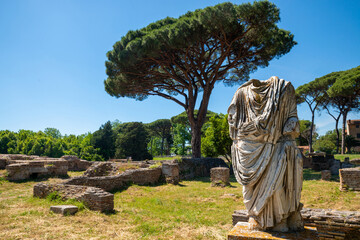 Ostia Antica, Rome Italy.Detail of a toga statue in the archaeological park site with brick architectural buildings from the imperial era, in summer with blue sky pine trees. Italy. - obrazy, fototapety, plakaty