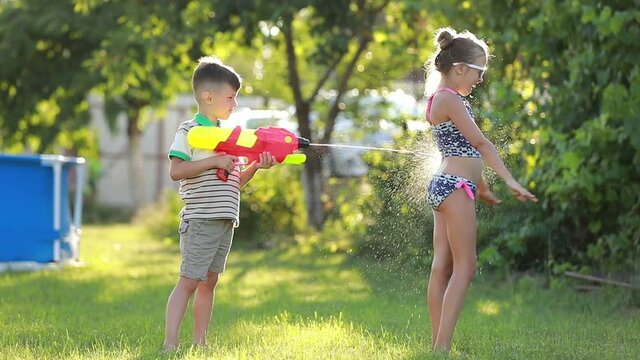 little boy is Splashing water on her older sister. Slow motion video of happy cheerful kids having fun in garden with water guns.