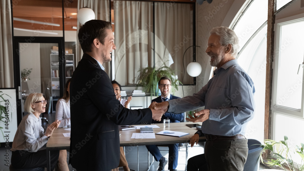 Poster Happy older male team leader giving handshake to excited employee, welcoming new team member, congratulating on good work, hiring for job, career promotion. Team clapping hands and laughing