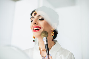 cheerful indian bride applying face powder with cosmetic brush on white