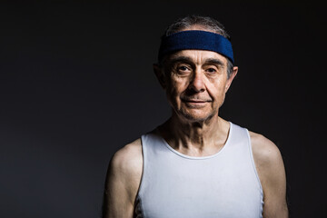 Fototapeta premium Elderly athlete wearing white sleeveless shirt, with sun marks on the arms, and blue headband, on a dark background. Sports and victory concept.