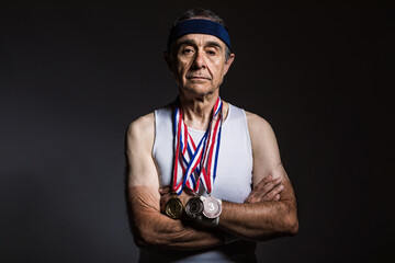 Elderly athlete wearing white sleeveless shirt, with sun marks on his arms, with three medals on his neck, with his arms crossed, on a dark background. Sports and victory concept.