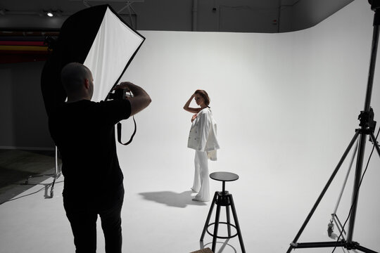 Fashion photography in a photo studio. Professional male photographer taking pictures of beautiful woman model on camera, backstage