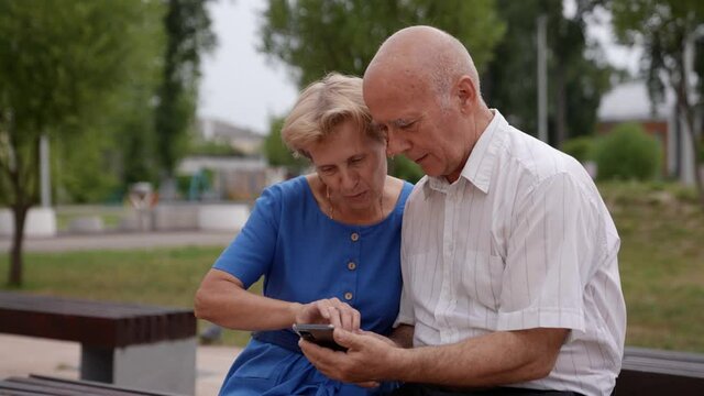 An Elderly Married Couple Enjoys Using A Mobile Application In The Park, Holding A Smartphone, Looking At The Mobile Phone Screen, Browsing Social Networks