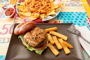 beef burger with lots of stewed onions, toast, iceberg lettuce, melted cheese and tomato served with french fries at a restaurant table with chili con carne nachos
