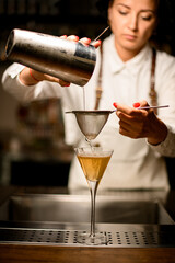female bartender pouring cocktail from a shaker through the sieve to glass