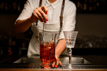 view of female bartender gently stirring with spoon cocktail in transparent mixing cup