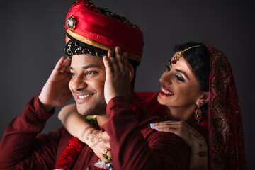 cheerful indian bride hugging bridegroom in turban isolated on grey