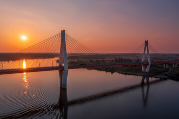 Aerial view of a white suspension bridge with two huge pillars above a river at sunrise in a rural area