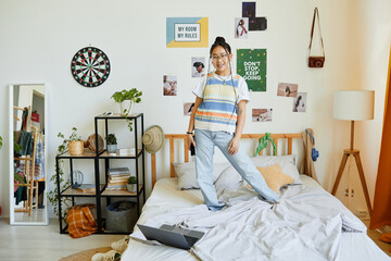 Full length portrait of teenage Asian girl standing on bed in cozy room interior and looking at...