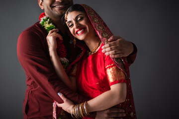 Indian man in turban hugging happy bride in traditional headscarf isolated on grey