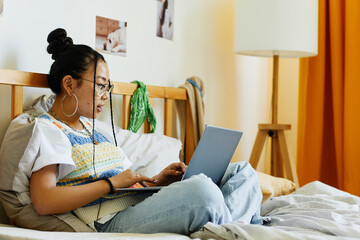 Side view portrait of teenage Asian girl using laptop while sitting on bed in cozy room interior,...