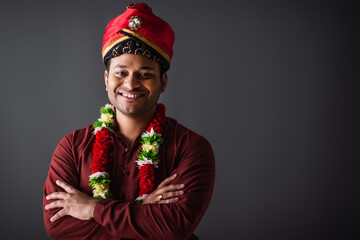 smiling indian man in turban and floral garland standing with crossed arms isolated on grey