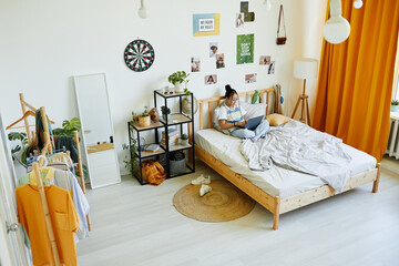 Wide angle portrait of teenage Asian girl using laptop while sitting on bed in cozy room interior,...