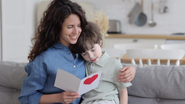 Mother day celebration: caring mom embrace cute son greeting mommy with holiday hold heart postcard kiss small boy in temple. Kid congratulating mom with birthday. Family tenderness and love concept