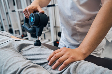 Faceless physical therapist with massage percussion gun treats girls leg close-up. Muscle stimulator