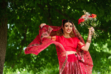 happy indian bride in red sari and traditional headscarf with ornament holding flowers