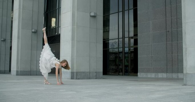 Young dancer in white dress does leg swing in slow motion outdoors, balerina dances in the gallery of columns architectural complex, 4k Prores HQ 120 fps