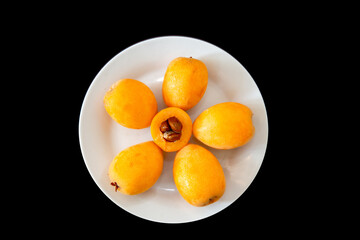 Ripe loquats on white plate on black background top view. One cut in half showing the inside of the fruit with the seeds ( Eriobotrya japonica )