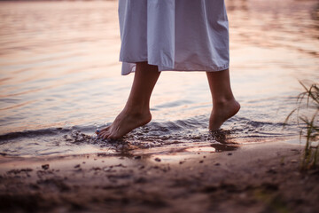 Women's feet in the river. Lifestyle close-up on the feet of a woman in a long white dress walking along the riverbank at sunset. 